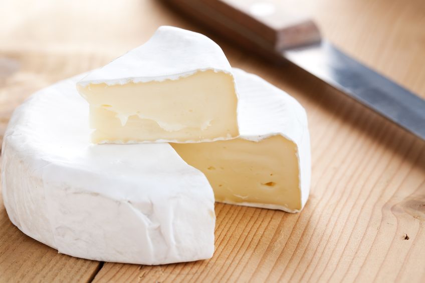 A picture of Brie Cheese on a cutting board. Behind the cheese to the right there is a small knife. There is a slice of cheese cut out. The slice is placed on top of the rest of the cheese.