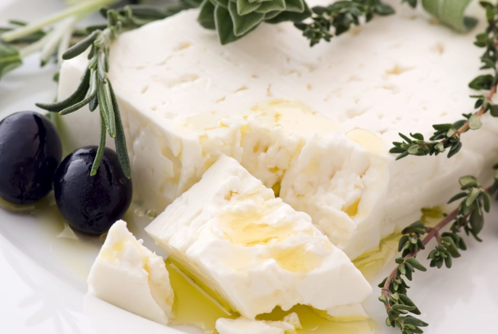 feta cheese on a plate with olive oil drizzled over it. There are black olives to the left and herbs on top. The corner of the feta is crumbled.