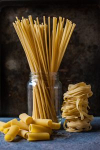 3 different types of on a table with a black background. Going left to right, there is penne on the table, spaghetti in a jar, and piles of linguine stacked beside the jar of pasta.