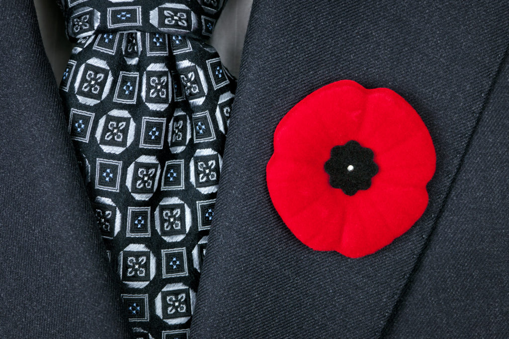 close up of a man dressed in a suite. All you can see is his lapel and tie. The suit is dark grey and his tie is printed in the colours of white, dark blue and light blue. To the right of his tie, on his lapel is a bright red poppy pin.