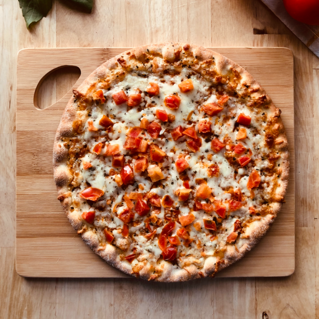 Image of Vince's Pizza on a cutting board. Around the board are tomatoes and basil. There is cheese and tomatoes on the pizza