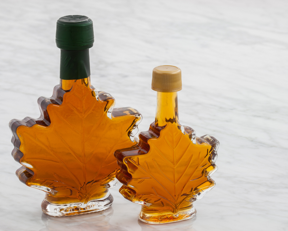 Image of 2 maple syrup bottles in the shape of a maple leaf. One bottle is slightly in front of the other. The back bottle has a black lid, the front one has a gold colour lid.
