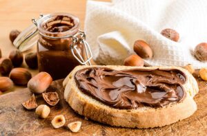Nutella spread onto a slice of toast beside a jar of Nutella and hazelnuts